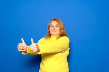 Caucasian woman in her 40s wearing a yellow sweater spreading her arms with her thumbs up in approval, isolated on blue studio background.
