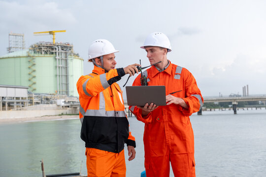 Two workers in orange safety suits communicate at a construction site near the water using a laptop and radio device