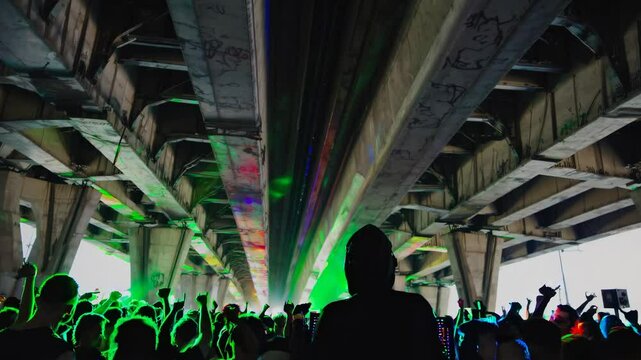 Silhouetted DJ and crowd under a bridge with colorful lights, captured from a low angle, creating a vibrant, immersive video party atmosphere.