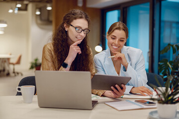 Two women are actively engaged in teamwork as they analyze data on a tablet within a contemporary...