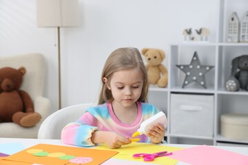 Cute little girl making handmade project with color paper and bottle of glue at table indoors