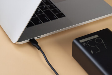 Charging laptop with power bank on beige background, close-up.
