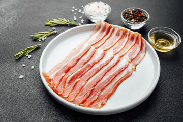Raw bacon slices on white plate with pepper, salt and rosemary