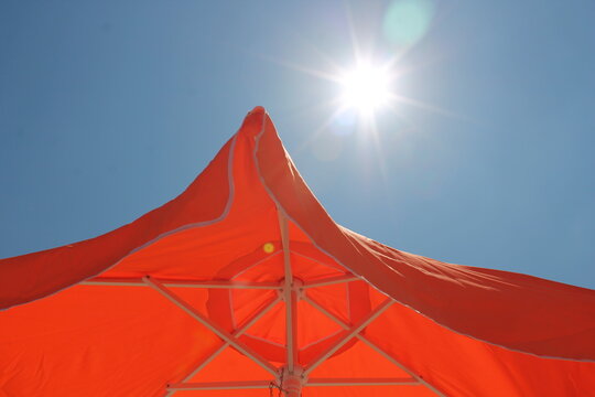upward view beneath a bright red circus or event tent canopy against a clear blue sky, with the sun shining through the fabric and supporting structure. The vibrant red fabric creates bold geometric 