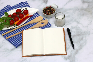Open notebook with blank pages for recipes with wooden spoon, spices and tomatoes.