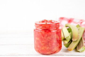 Pink and green rhubarb sweet jam. Homemade tasty sweet rhubarb confiture preserves, in small jar on white wooden table, with fresh rhubarb in the background