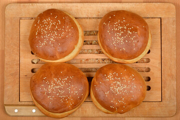 Four golden brown homemade burger buns, sprinkled with sesame seeds, are arranged on a wooden cutting board
