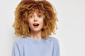 Surprised young woman with curly hair expressing excitement on a plain light background, wearing a...