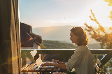 Young woman sitting on balcony with view of mountains and sunset working using laptop