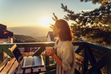 Young woman sitting on balcony with view of mountains and sunset working using laptop