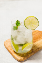 A refreshing cocktail served in a clear glass filled with ice cubes. The drink garnished with a lime slices and mint leaves. The glass is placed on a wooden board, set against a neutral background.