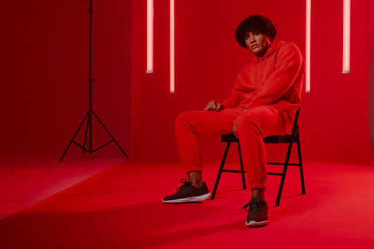 Sitting male wearing red tracksuit on black folding chair in studio, with LED tubes, copy space