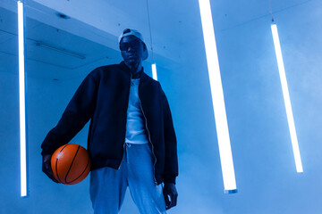 African American man standing in studio with blue LED lights and basketball in hand, copy space