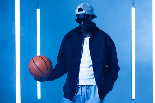 Standing African American man in sportswear, holding basketball in misty blue studio with LED tubes
