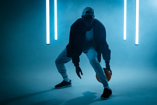 Dribbling African American man with backward cap crouching in hazy studio, with basketball and LEDs