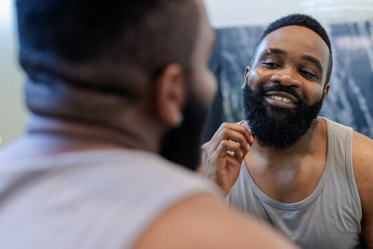 Smiling African American man stroking full beard at bathroom mirror with gray tank top