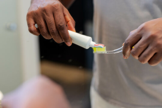 African American man squeezing toothpaste onto clear toothbrush at bathroom sink, with striped tube