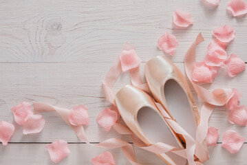 Fototapeta na wymiar Pastel pink ballet shoes background. New pointe shoes with satin ribbon and rose petals lay on white rustic shubby chic wood, top view with copy space, soft toning
