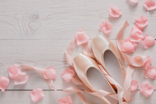 Pastel pink ballet shoes background. New pointe shoes with satin ribbon and rose petals lay on white rustic shubby chic wood, top view with copy space, soft toning