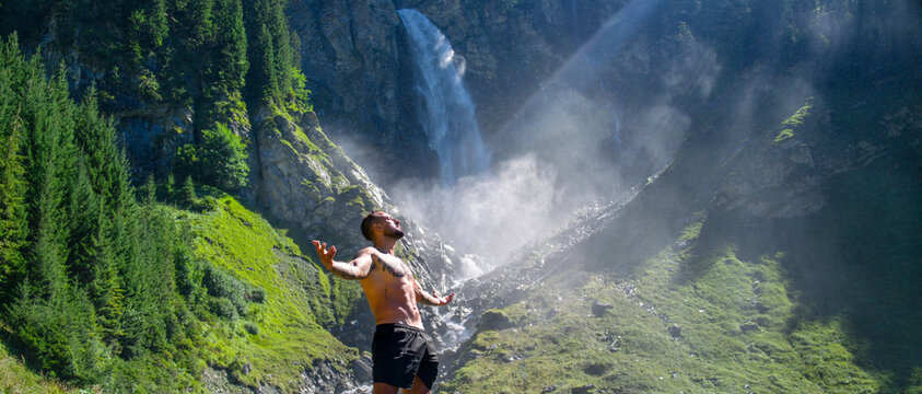 Carefree man in waterfall. Man freedom in waterfall landscape. Muscular man breathing fresh air on beauty waterfall. Topless model with raised hands on mountain nature landscape. Yoga by waterfall.