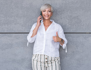 Yes, i am on the phone, happy trendy middle age woman talks on the smartphone and gesturing like sign, against gray background - wall. Pleased woman using the phone