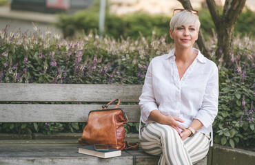Pretty blond adult woman sitting on bench in the city. Beautiful female person dressed in white summer shirt. Outdoors.