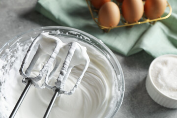 Hand mixer with whisks and bowl of whipped cream on grey textured table, closeup