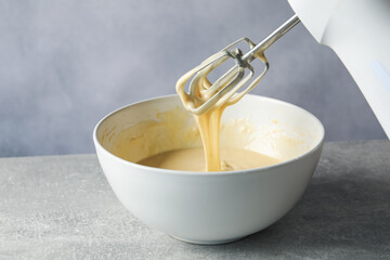 Hand mixer with whisks and bowl of dough on grey textured table, closeup