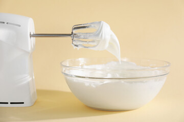 Hand mixer with whisks and bowl of whipped cream on beige background, closeup
