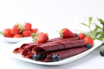 Tasty fruit leather rolls, strawberries and blueberries on white table, closeup. Space for text