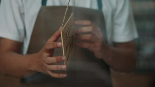 Closeup young waiter hands turn entrance sign Closed to Open on glass cafe door. Fast food restaurant worker in apron shows inviting plate. Local coffee shop customer service