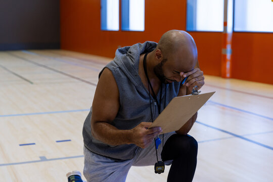 Mid-adult male coach kneeling on indoor basketball court, wiping sweat with clipboard and stopwatch - Powered by Adobe