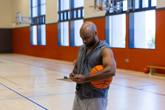 male coach standing on indoor basketball court, holding basketball and noting on clipboard with pen