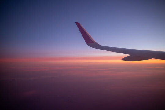 An airplane wing is captured against a stunning twilight sky filled with a gradient of blue purple orange and pink, symbolizing air travel and the serene beauty of a sunset from above the clouds.