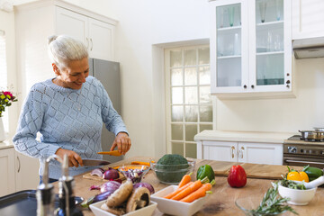 Senior woman chopping carrot in modern home kitchen, with knife on wooden board and vegetables