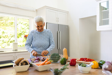 Senior woman slicing vegetables on wooden island in home kitchen, with cutting board, chef's knife