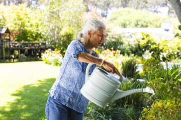 Senior woman watering flowering plants in residential garden, with mint-green watering can © wavebreak3