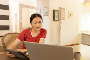 Young Asian woman leaning at wooden table in home, using laptop and tablet with coffee mug