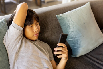 Young Asian man reclining on gray couch at home with teal pillows, using smartphone, copy space