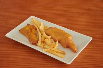 A plate of fried fish and french fries. The fish is cut in half and the fries are cut into small pieces