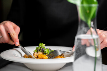 A person enjoys a plate of pasta with fresh greens, using a fork and knife. The elegant setting...