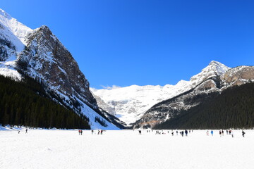 Obraz premium Lake Louise, Alberta, Canada - Apr 13, 2025: Tourists walking on the frozen Lake Louise. Beautiful Lake Louise in spring. The lake is frozen with unreal, scenic nature views in popular tourism area.