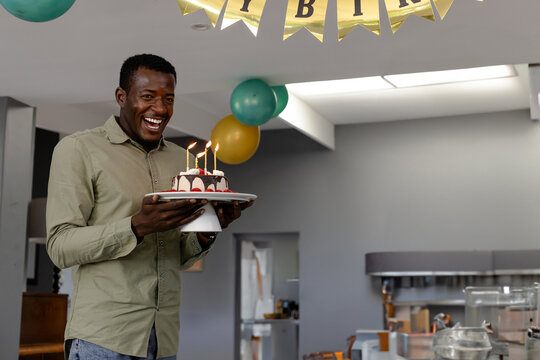 African American man holding cake with lit candles in kitchen, with balloons, banner, copy space