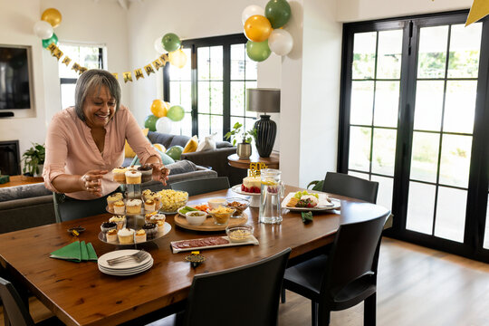 Senior woman arranging cupcakes on tiered stand in dining room at home, with birthday banner