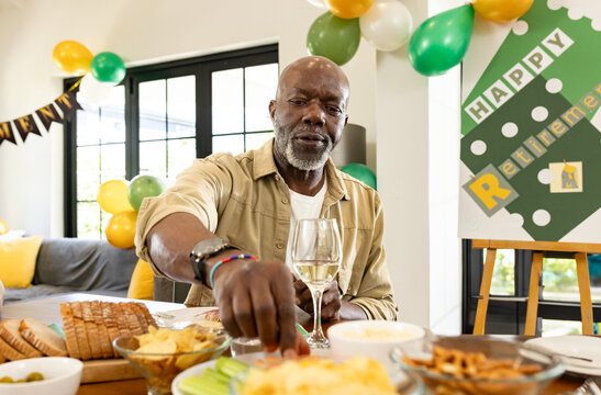 Senior African American man grabbing snack from table at retirement celebration, holding wine glass