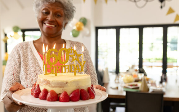 Smiling senior woman holding 60th birthday cake in dining room, balloons and bunting, copy space