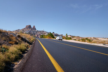 A road with a distinctive yellow line leads to a historical hill town in Cappadocia, Turkije bathed in sunlight under a clear, expansive sky evoking wanderlust and adventure.