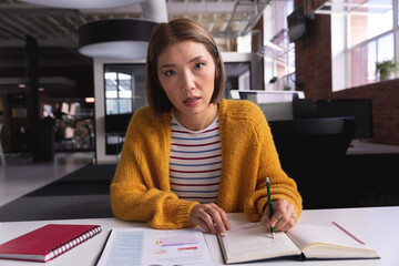 Mid-adult Asian woman writing notes at desk in modern office, with green pen and printed charts