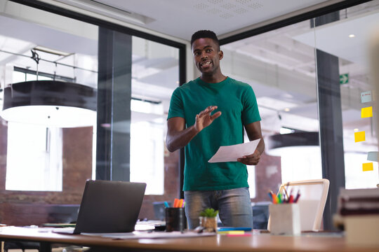 African American man presenting document in modern open-plan office, holding paper and laptop