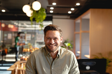 Mid adult man smiling at camera seated in modern cafe amid wooden decor and lush plants